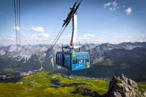 Eine blaue Seilbahn fährt über eine grüne Berglandschaft mit felsigen Gipfeln und einem Tal darunter unter einem teilweise bewölkten Himmel.
