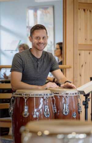 Ein Mann in einem grauen Hemd sitzt an einer hölzernen Conga-Trommel und lächelt in die Kamera, während im Hintergrund Menschen und Notenständer zu sehen sind.